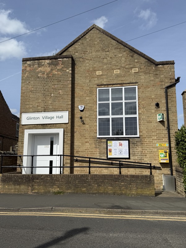 Glinton Village Hall seen from the road, with its name sign and noticeboard beside the entrance